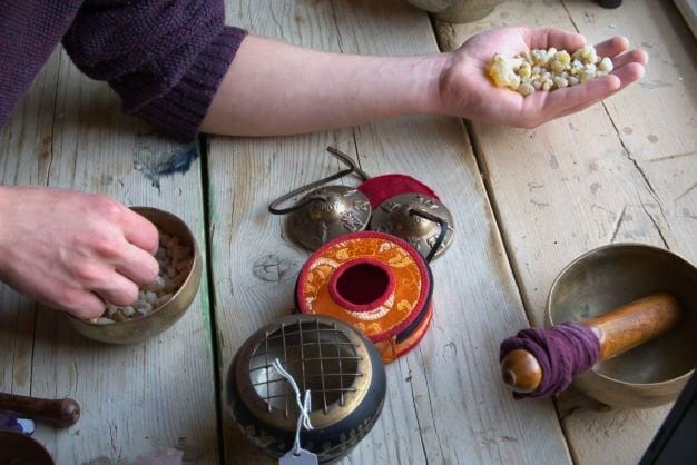 May include: A set of Tibetan singing bowls, a small incense burner, and a hand holding a pile of resin. The singing bowls are made of brass and have a red and orange fabric covering. The incense burner is made of brass and has a black and gold finish. The resin is a light brown colour.