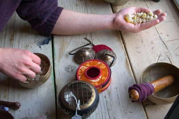 May include: A set of Tibetan singing bowls, a small incense burner, and a hand holding a pile of resin. The singing bowls are made of brass and have a red and orange fabric covering. The incense burner is made of brass and has a black and gold finish. The resin is a light brown colour.